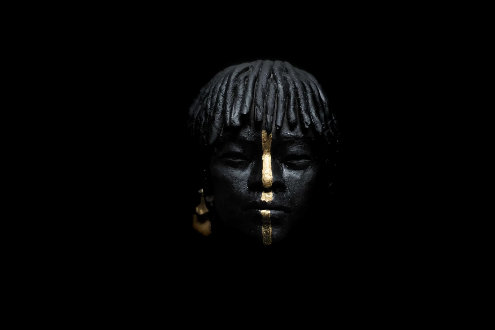 Photographie de sculpture sur fond noir : visage marqué d'une ligne de feuille d'or, Terres Ethniques