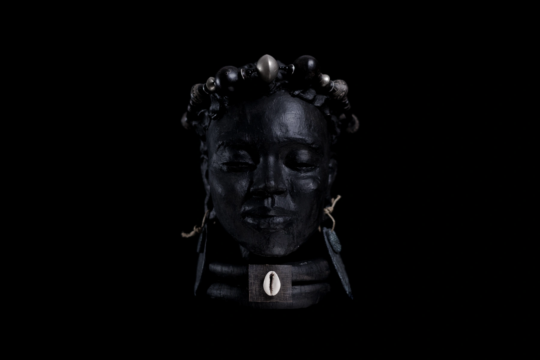 Sculpture de visage portant un bandeau de perles et un cauri, photographiée sur fond noir